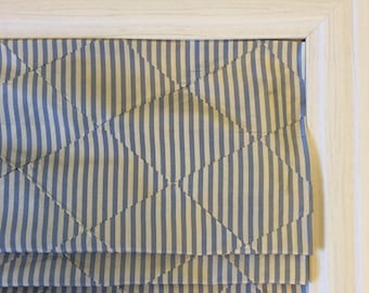 May include: A white window frame with a blue and white striped fabric roman shade. The shade is folded up and has a diamond pattern.
