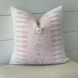 Op de afbeelding: Een wit kussen met een geometrisch patroon in roze en groen. Het kussen ligt op een lichtbeige kussen.