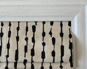 May include: A white window frame with a black and white patterned roman shade. The shade has a geometric pattern of vertical lines with black dots.