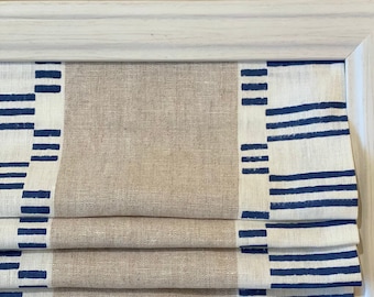 May include: A white and beige roman shade with blue stripes along the edges. The shade is made of a linen fabric and has a relaxed, casual look.