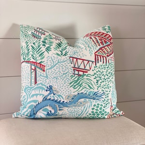 Könnte beinhalten: Dekoratives quadratisches Kissen mit einem blauen Drachendesign. Das Kissen hat einen weißen Hintergrund mit einem Muster aus roten Strukturen, grünem Laub und einem blauen Drachen. Es liegt auf einer beige gepolsterten Oberfläche.