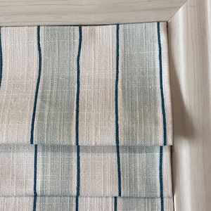 May include: Close-up of a white and blue striped roman shade with a textured fabric. The shade is partially raised, showing the layers of fabric.