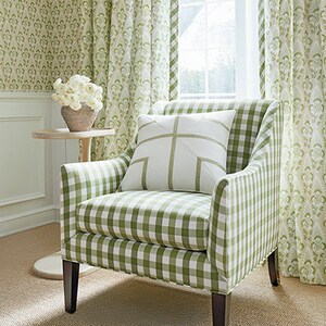 May include: A green and white gingham armchair with a white pillow with a green cross design. The chair is in front of a window with green patterned curtains.