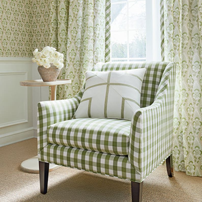 May include: A green and white gingham armchair with a white pillow with a green cross design. The chair is in front of a window with green patterned curtains.
