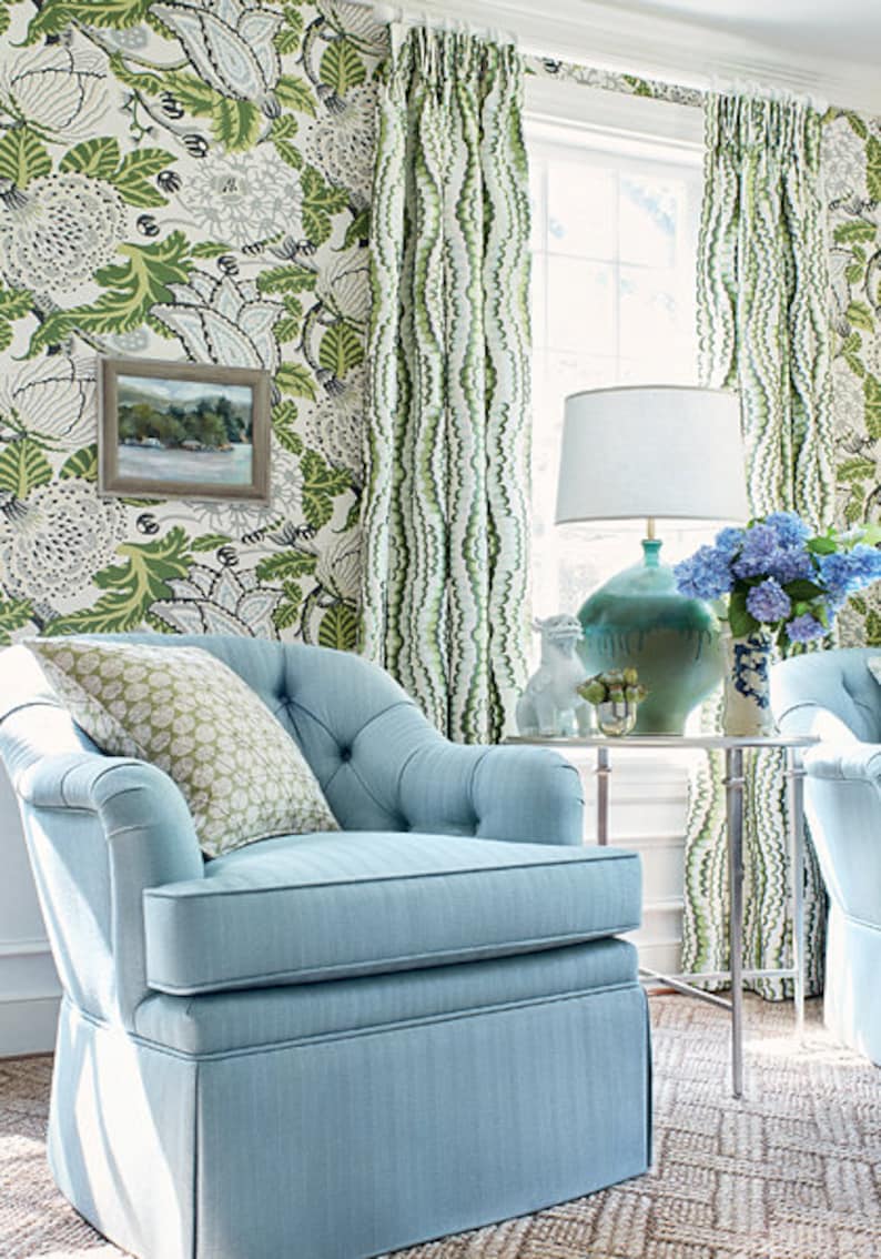 Pu&ograve; includere: Una poltrona blu chiaro con schienale capitonn&eacute; e un cuscino con motivo floreale. La poltrona &egrave; davanti a una finestra con tende bianche con motivo verde e bianco. Un tavolo con una lampada e un vaso di fiori si trova accanto alla poltrona.