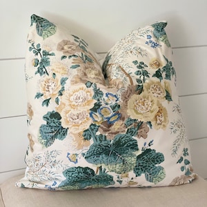 May include: A decorative throw pillow with a floral pattern. The pillow features large, cream-colored flowers with green and blue foliage against a cream background. The pillow is placed on a neutral-toned upholstered bench.