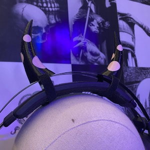 May include: Black headband with two pink and white devil horns with polka dots. The headband is on a white mannequin head.