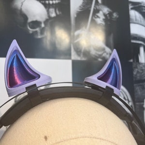 Puede incluir: Un par de orejas de gato en una diadema negra. Las orejas son de color lavanda con un degradado de azul, morado y rosa en el interior. La diadema es negra y parece estar hecha de plástico o un material similar.