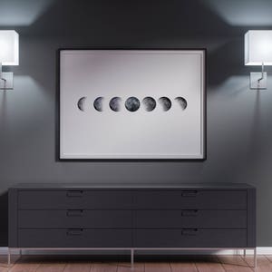 Op de afbeelding: Een zwarte commode met zes laden staat voor een grijze muur. Een ingelijst afdruk van de maanfasen hangt boven de commode. Twee vierkante wandlampen met witte lampenkappen verlichten de kamer.