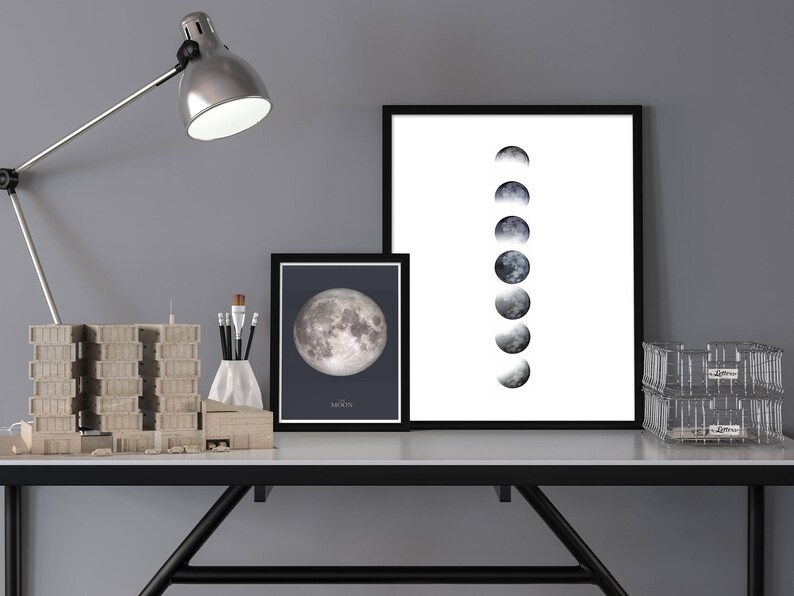 Op de afbeelding: Twee zwart ingelijste prints van de maanfasen op een witte achtergrond. De print aan de linkerkant toont een volle maan, terwijl de print aan de rechterkant een reeks maanfasen toont. De prints staan op een bureau met een lamp, een modelgebouw en een draadmand.