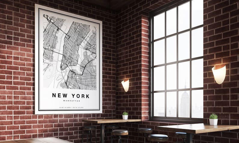 May include: A framed black and white map of New York City, Manhattan, hangs on a red brick wall. The map is labeled "NEW YORK" with coordinates. The room features a large window, wooden tables, and decorative lighting.