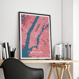 May include: Framed New York City map print in a modern style. The map features a red and blue color scheme, with the city's streets and waterways clearly defined. The text "NEW YORK MANHATTAN" is displayed at the bottom of the map. The print is displayed on a wooden desk.