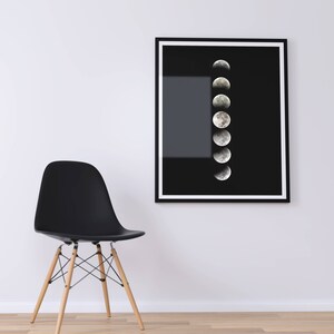 May include: A black framed print of a moon phases illustration on a white wall with a black chair in the foreground.