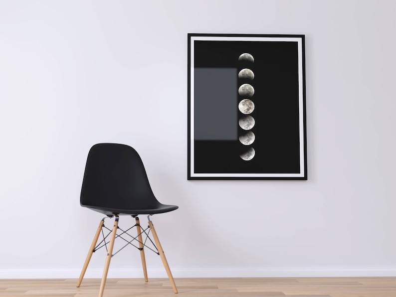 May include: A black framed print of a moon phases illustration on a white wall with a black chair in the foreground.