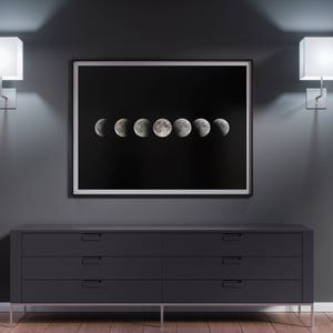 May include: A black and white framed print of the moon in different phases hanging on a dark gray wall above a dark gray dresser with four drawers. Two white square wall sconces are on either side of the print.
