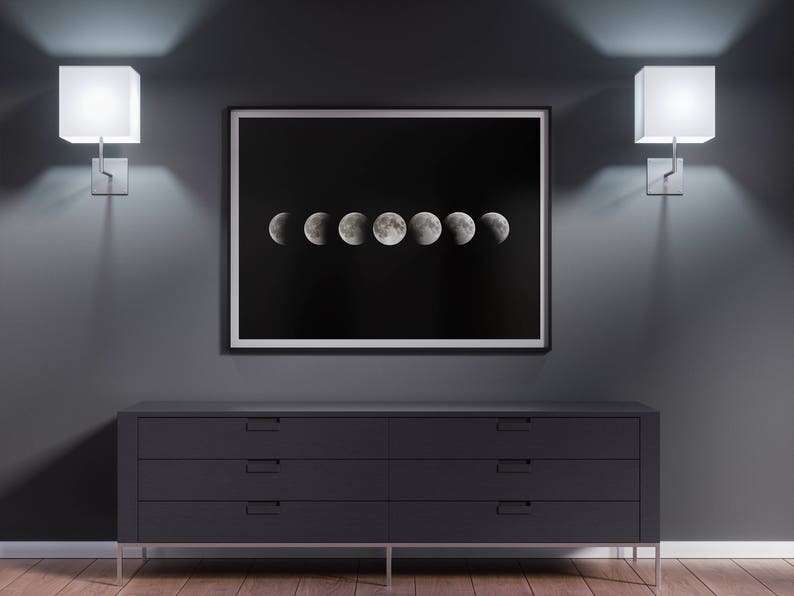 May include: A black and white framed print of the moon in different phases hanging on a dark gray wall above a dark gray dresser with four drawers. Two white square wall sconces are on either side of the print.