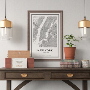 May include: A framed black and white map of New York City, labeled "NEW YORK MANHATTAN," sits on a dark wood table. The table also holds a wooden box, a terracotta pot with a plant, books, and a candle. Two gold and glass pendant lights hang above.