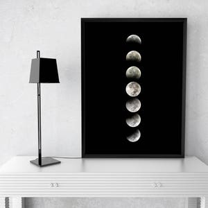 May include: A black framed print of a moon in various phases of a lunar cycle against a black background. The print is displayed on a white table with a black lamp.