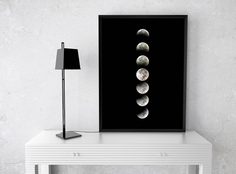 May include: A black framed print of a moon in various phases of a lunar cycle against a black background. The print is displayed on a white table with a black lamp.
