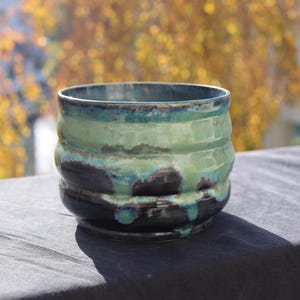 May include: A handmade ceramic bowl with a unique, textured design. The bowl features a combination of green, teal, and black colors, with a glossy finish. The exterior has a ribbed pattern, adding visual interest. The bowl is suitable for serving or display.