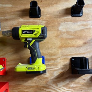 May include: Two Ryobi power tools mounted on a wall with black plastic holders. The tools are yellow and black with the Ryobi logo and the text "ONE+" on the side. The tools are mounted on a wooden wall.