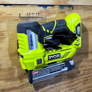 May include: A neon green and black Ryobi AirStrike cordless nail gun. The tool has a black handle and a silver magazine. The brand name and model are visible on the side. The nail gun is resting on a wooden surface.