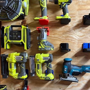 May include: A wall-mounted tool organizer with various power tools, including a Ryobi air compressor, a Ryobi router, a Ryobi nail gun, a Ryobi brad nailer, a Makita jigsaw, and a Ryobi fan. The tools are mounted on black and blue plastic brackets.