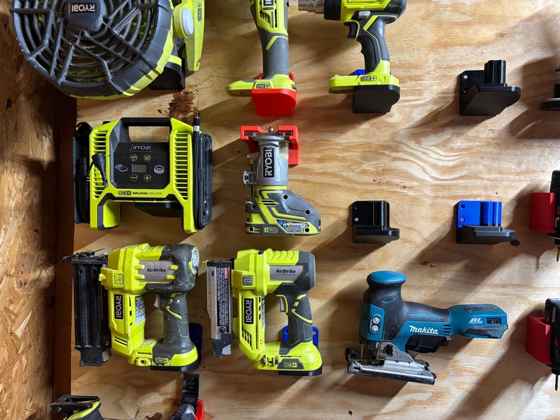 May include: A wall-mounted tool organizer with various power tools, including a Ryobi air compressor, a Ryobi router, a Ryobi nail gun, a Ryobi brad nailer, a Makita jigsaw, and a Ryobi fan. The tools are mounted on black and blue plastic brackets.