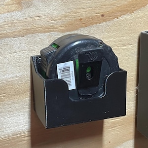 May include: A black tape measure in a black holder mounted on a wooden surface. The tape measure has a green accent and a barcode sticker. The holder is square and has a minimalist design. The image is well-lit and shows the details of the tape measure.