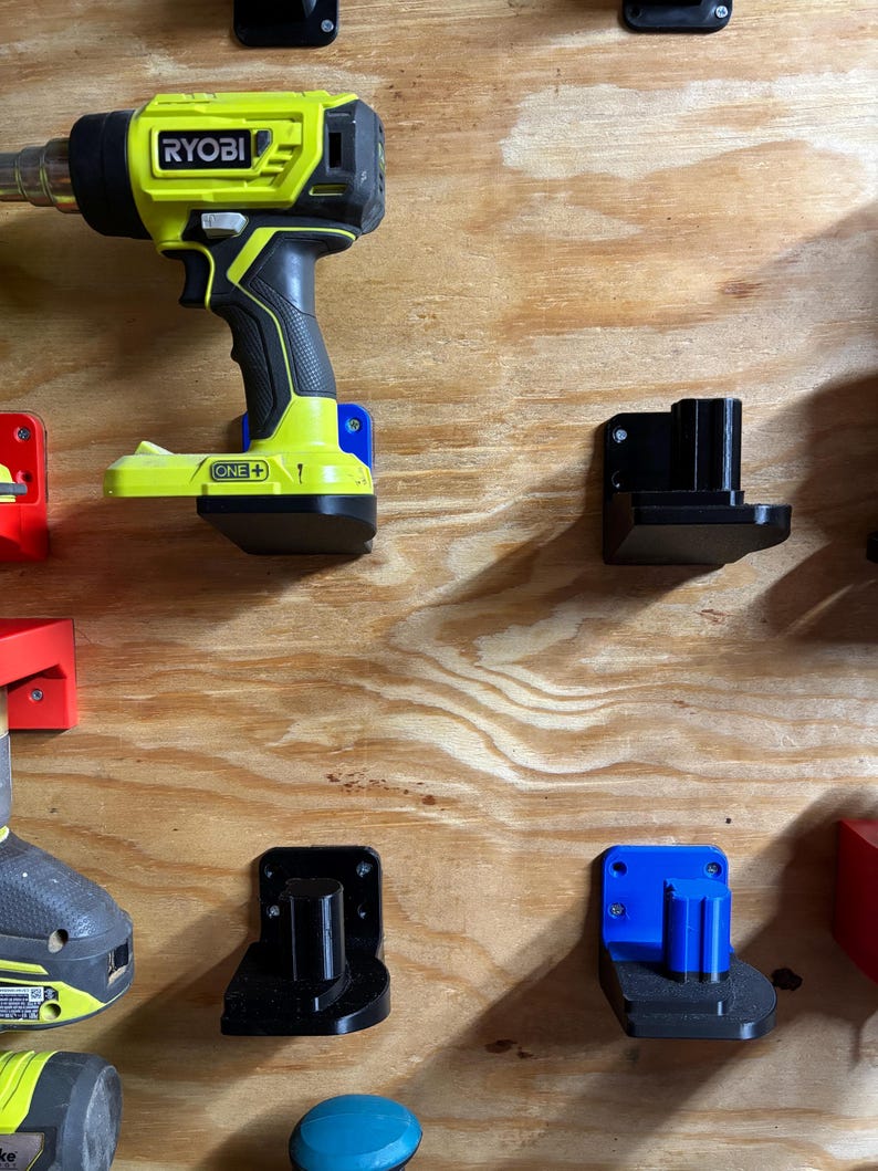 May include: A yellow and black Ryobi drill with a black and blue wall mount. The drill is labeled "RYOBI ONE+". There are three other wall mounts, one black and two blue, on a wood panel.