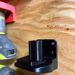 May include: A black plastic wall mount for a power tool. The mount has a round hole in the center and a curved shape on the bottom.