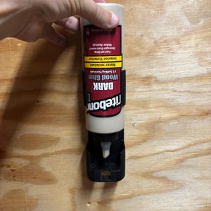 May include: A bottle of Titebond DARK Wood Glue, held by a hand. The label indicates it is water-resistant and suitable for interior and exterior use. The bottle is beige with a black label and a black dispenser.