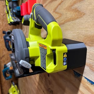 May include: A lime green and black circular saw with a black handle and blade guard. The saw has the brand name "RYOBI" on the blade guard. Other power tools are visible in the background.