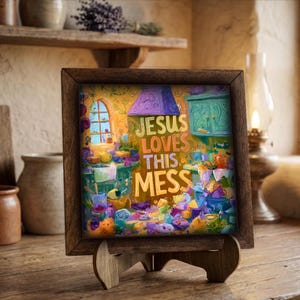 May include: A framed art print with the text "JESUS LOVES THIS MESS" in colorful lettering. The artwork depicts a vibrant, whimsical kitchen scene with various objects and a window. The frame is made of dark wood and is displayed on a wooden easel.