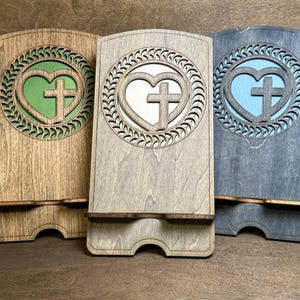 May include: Three wooden phone stands with a heart and cross design. The stands are in natural wood, gray, and blue.