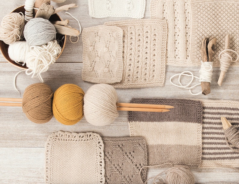 May include: An overhead shot of various knitting supplies. Several skeins of yarn in shades of white, gray, brown, and yellow are arranged with wooden knitting needles and knitted swatches. A wooden bowl holds more yarn and driftwood.