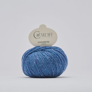 May include: A ball of blue Cardiff Cashmere Classic yarn with white flecks. A light-colored tag above the yarn displays the brand name and the word "Cashmere". The yarn is a soft, textured material, perfect for knitting or crocheting projects.