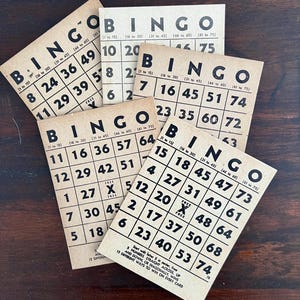 May include: A collection of vintage bingo cards with black numbers and the word "BINGO" printed in black on a tan background. The cards are arranged on a dark wooden surface, with some overlapping.