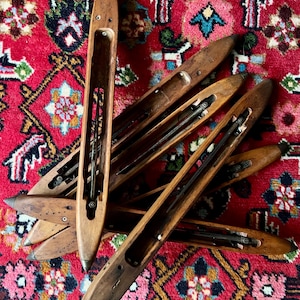 May include: A collection of vintage wooden weaving shuttles, each with a long, boat-shaped body and a central cavity. The shuttles are a warm brown color and rest on a vibrant red rug with a detailed floral pattern.