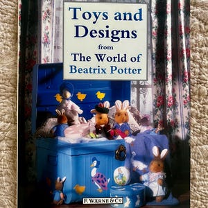 May include: A hardcover book titled Toys and Designs from The World of Beatrix Potter, featuring a blue storage chest filled with plush rabbit and duck toys, a small round box, and a blue knitted blanket, with F. Warne & Co printed at the bottom.