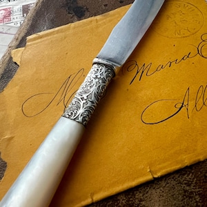May include: A vintage butter knife with a mother-of-pearl handle and ornate silver detailing. The knife rests on a yellowed envelope with handwritten script. The blade is silver and the handle is a creamy white color.