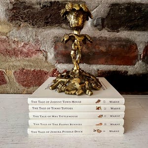 May include: A golden brass candlestick with a detailed floral design sits atop a stack of five books. The books are titled "The Tale of Johnny Town-Mouse", "The Tale of Timmy Tiptoes", "The Tale of Mrs. Tittlemouse", "The Tale of the Flopsy Bunnies", and "The Tale of Jemima Puddle-Duck".