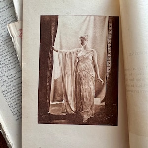 May include: A black and white photograph of a woman in a long white dress standing in a doorway. The woman is holding the edge of a curtain with her left hand and her right hand is at her side. The doorway is decorated with a geometric pattern.