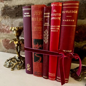 May include: A collection of antique books bound in shades of red and burgundy, tied together with a velvet ribbon. The spines display gold lettering and titles such as "British Orations" and "Just His Luck". A decorative brass candlestick is in the background.