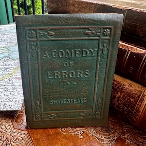 May include: A green hardcover book titled A Comedy of Errors by Shakespeare. The front cover features an embossed rectangular border with floral motifs at the corners and sides. The text is centered in a dark green font on the textured cover.