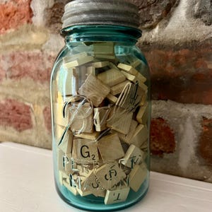 May include: A vintage blue-green glass mason jar filled with wooden Scrabble tiles. The jar has a metal lid and is set against a brick wall background. Some tiles are visible with letters and point values. The jar is sitting on a white surface.
