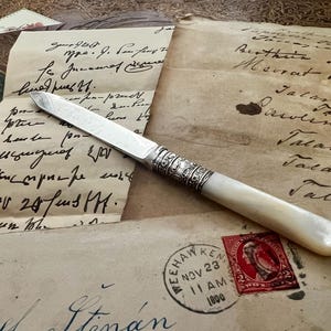 May include: An antique letter opener with a silver blade and a white handle. The handle has ornate silver detailing. The opener rests on aged paper with handwritten script and a vintage stamp.