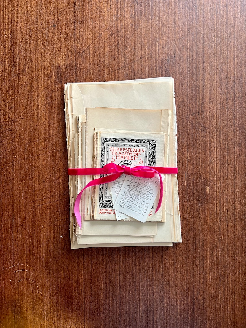May include: A stack of vintage paperbacks tied together with a pink ribbon. The top book has the title "Shakespeare's Tragedy of Hamlet" printed on the cover.