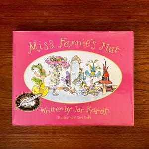 May include: A pink hardcover book titled "Miss Fannie's Hat" with illustrations of hats and characters. The book is written by Jan Karon and illustrated by Toni Goffe. An autographed copy seal is on the lower left.