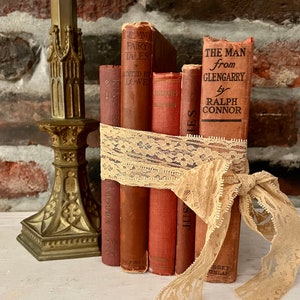 May include: A stack of vintage hardcover books with brown and red covers, tied together with a beige lace ribbon. The books have titles like "Grammy Fairy Tales", "The Man from Glengarry", and "Sunset Island".